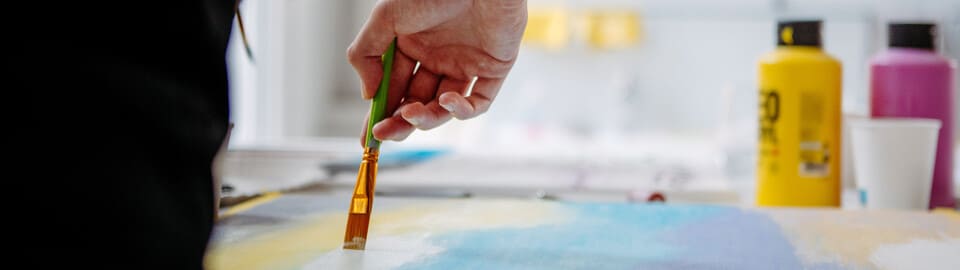 Eine Hand hält einen Pinsel und streicht mit weißer Farbe über ein Blatt Papier, das mit etwas blauer Farbe bemalt ist.